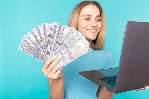 Portret podekscytowanej zadowolonej dziewczyny trzymającej banknoty z laptopem na niebieskim tle. Portret uśmiechniętej młodej kobiety trzymającej w rękach banknoty i laptop — Zdjęcie stockowe