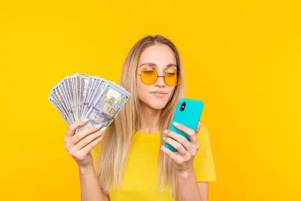Portrait of a young blonde woman holding bunch of money banknotes and looking at mobile phone over yellow background — Stock Photo, Image