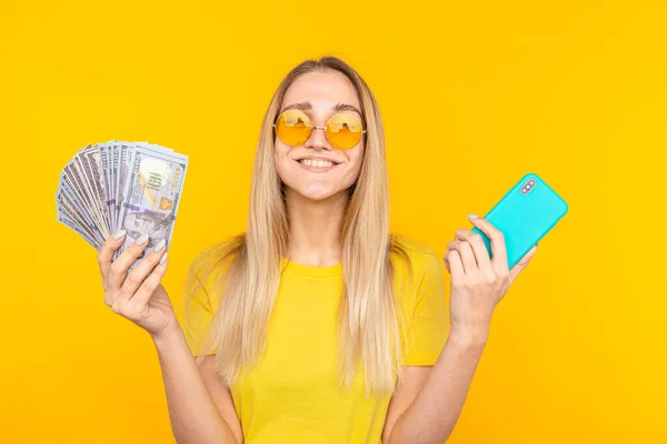 Portret szczęśliwej młodej kobiety trzymającej kupę banknotów i pokazującej telefon komórkowy na żółtym tle — Zdjęcie stockowe