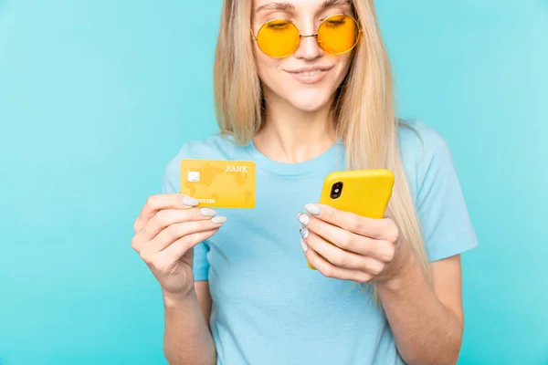 Image of young woman holding cell phone and credit card in hands isolated over blue background