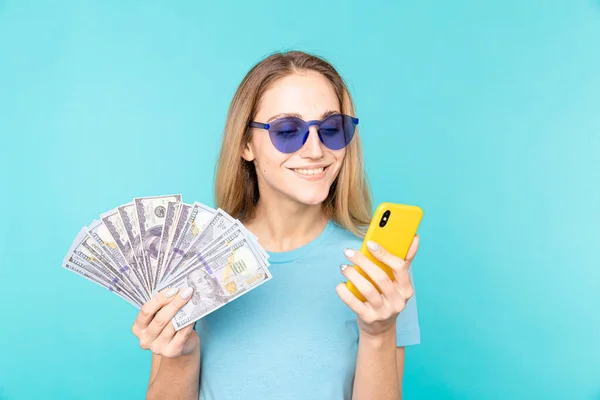 Uśmiechnięta młoda blondynka dziewczyna w ciemnych okularach trzyma gotówkę w banknotach dolarowych i smartfonie odizolowanym na niebieskim tle w studio — Zdjęcie stockowe