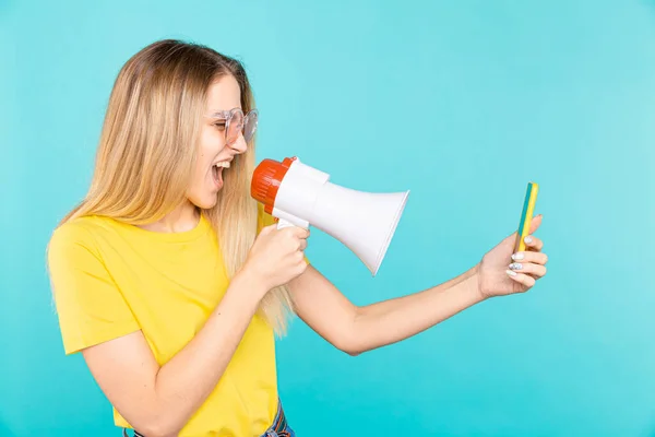 Boos jonge vrouw schreeuwen met megafoon naar mobiele telefoon op blauwe achtergrond — Stockfoto