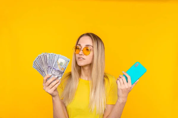 Young blonde woman holding mobile phone and looking at bunch of money banknotes over yellow background — Stock Photo, Image