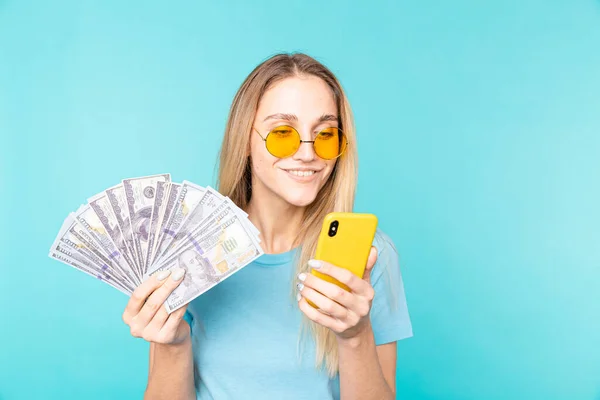 Obraz młodej uśmiechniętej kobiety trzymającej telefon komórkowy i banknoty pieniężne odizolowane na niebieskim tle — Zdjęcie stockowe