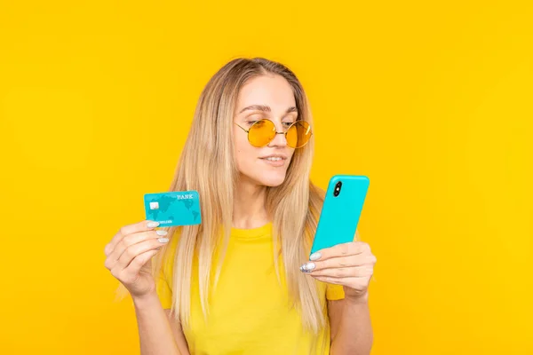 Portret van een jong blond meisje in een zonnebril met plastic creditcard tijdens het gebruik van de mobiele telefoon — Stockfoto
