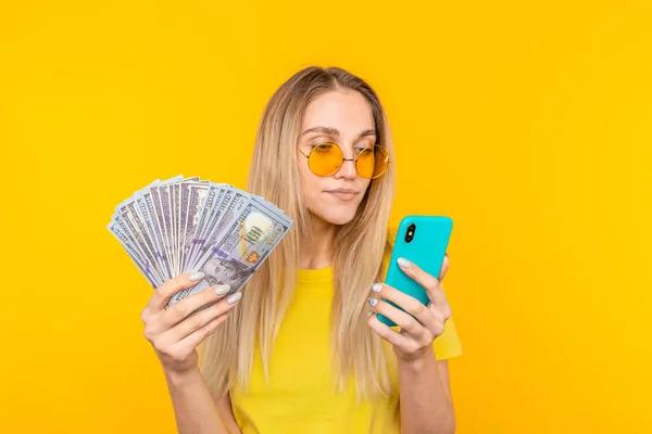 Portret młodej blondynki trzymającej kupę banknotów i patrzącej na telefon na żółtym tle — Zdjęcie stockowe