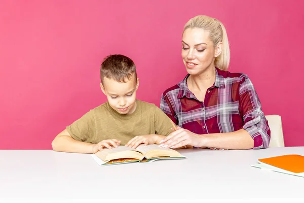 魅力的な若い女性と彼女の小さな息子がテーブルに座って一緒に宿題をしています。母は学校の授業で息子を助ける — ストック写真
