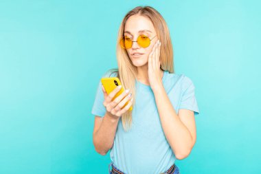Sarı güneş gözlüklü genç kadın mavi arka planda endişeli ve bir mesaj okuyor.