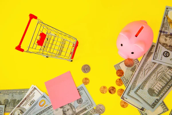 Winkelwagen en roze spaarvarken bank op een gele achtergrond, ruimte voor tekst. Financiën, sparen, crisis. Besparingen voor bedrijven of pensioenen — Stockfoto