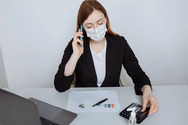 Wanita bisnis muda mengenakan masker wajah berbicara di telepon pintar duduk di meja tempat kerja di ruang kantor melindungi dirinya dari grippe Stok Foto