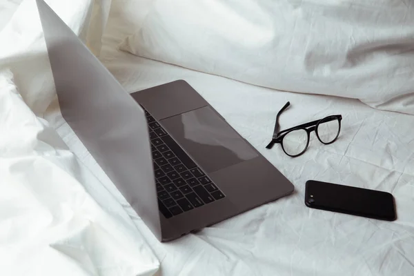 Ноутбук, очки и смартфон в постели на чистом белом белье. Работа на дому — стоковое фото