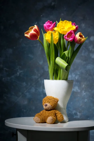 gri-mavi arka plan ve bir küçük peluş boz ayı yavrusu üzerinde beyaz bir vazoda sarı ve kırmızı laleler