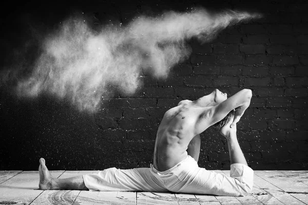 Um homem fazendo ioga em uma nuvem branca de poeira em uma sala escura. O conceito de energia. Foto em preto e branco. Eka pada rajakapo de hanumanasana. pose de pombo — Fotografia de Stock