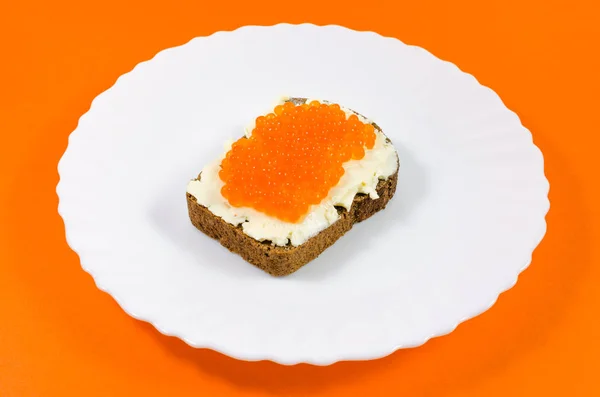 A piece of black rye bread on a white plate smeared with butter and red caviar — Stock Photo, Image