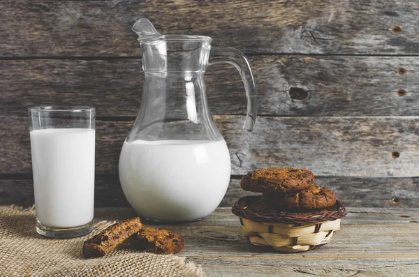Biscoitos de chocolate de aveia, jarro e copo de leite, fundo de madeira rústico . — Fotografia de Stock