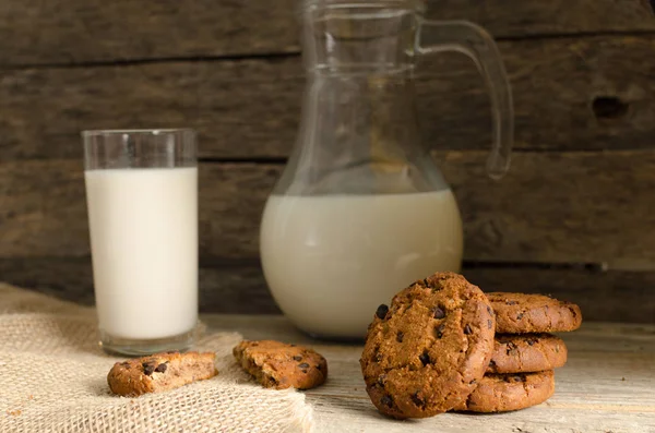 Biscoitos de chocolate de aveia, jarro e copo de leite, fundo de madeira rústico . — Fotografia de Stock