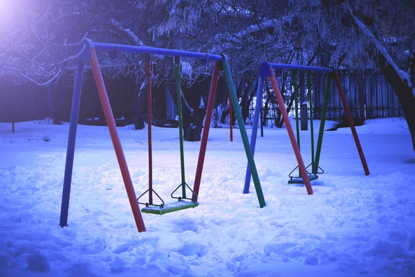 Детская площадка с качелями зимой в ночное время в свете уличного фонаря . — стоковое фото