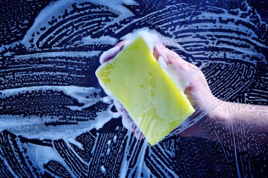 Man hand washes the glass covered with foam, yellow sponge on a dark background.