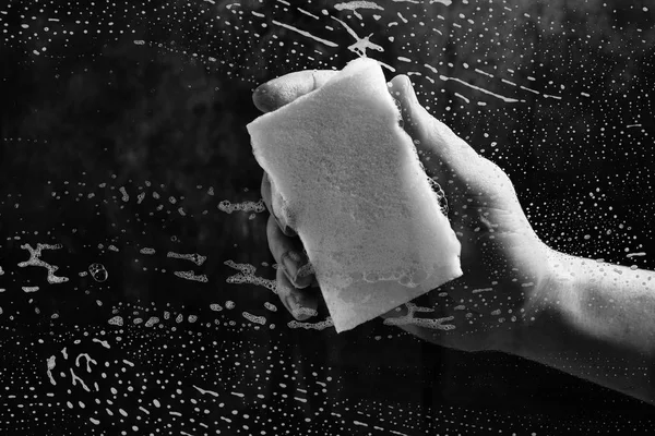 Tangan manusia mencuci kaca yang ditutupi dengan busa, spons kuning dengan latar belakang gelap . — Stok Foto