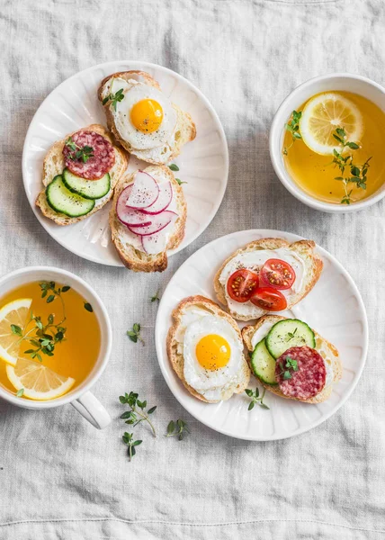 Delicious Breakfast - mini sandwiches with cream cheese, vegetables, quail eggs, salami and green tea with lemon and thyme. Sandwiches with cheese, cucumber, tomatoes, radish, thyme, lemon zest, fried quail eggs on a light background, top view. — Stock Image Delicious Breakfast - mini sandwiches with cream cheese, vegetables, quail eggs, salami and green tea with lemon and thyme. Sandwiches with cheese, cucumber, tomatoes, radish, thyme, lemon zest, fried quail eggs on a light background, top view. Stock Photo