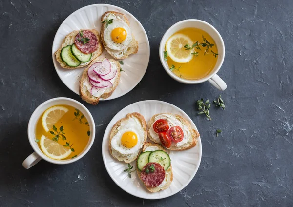 Mini sandwiches with cream cheese, vegetables, quail eggs, salami and green tea with lemon and thyme. Sandwiches with cheese, cucumber, tomatoes, radish, thyme, lemon zest, fried quail eggs on a dark background, top view. Delicious breakfast or snack — Stock Image Mini sandwiches with cream cheese, vegetables, quail eggs, salami and green tea with lemon and thyme. Sandwiches with cheese, cucumber, tomatoes, radish, thyme, lemon zest, fried quail eggs on a dark background, top view. Delicious breakfast or snack Stock Image