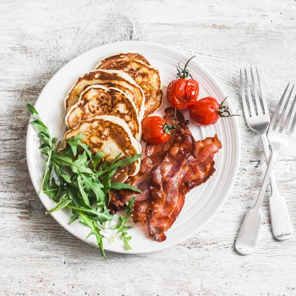 Traditional american breakfast - crispy bacon, pancakes with maple syrup, roasted tomatoes, arugula. On a light background, top view
