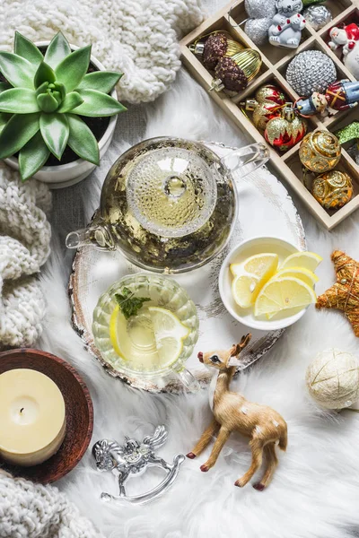 Glass teapot with green tea, flower, candle, knitted plaid and box of christmas decorations on a fluffy carpet on a light background. Christmas background — Stock Image Glass Teapot Green Tea Flower Candle Knitted Plaid Box Christmas Royalty Free Stock Photos
