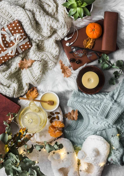 Cozy winter autumn things. Home coziness still life - soft slippers, knitted sweater, plaid, green tea with honey, tangerines, dry leaves, books, garland, gift box on a light background, top view. Autumn and winter relax mood. Flat lay