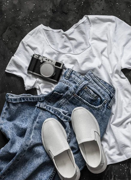 Simple women\'s look - white t-shirt, mom\'s jeans, white sneakers and a vintage camera on a dark background, top view. Fashion concept, flat lay