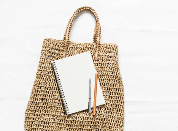 Eco bag made of straw and a clean notepad on a white background, top view. Shopping planning concept