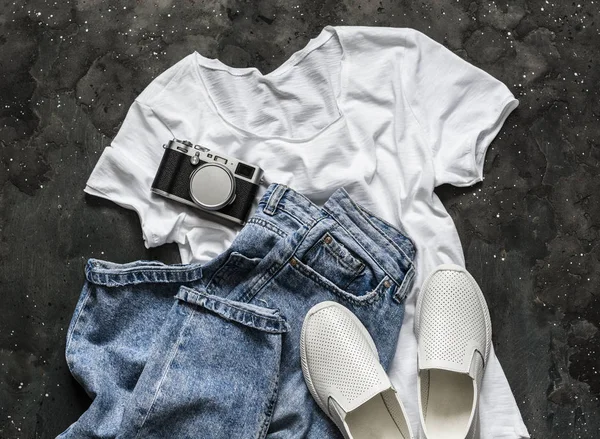 Simple women\'s look - white t-shirt, mom\'s jeans, white sneakers and a vintage camera on a dark background, top view. Fashion concept, flat lay