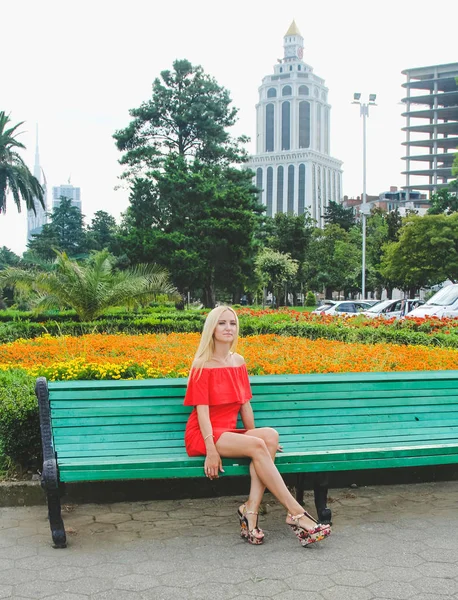 Элегантная блондинка в красном платье сидит на зеленой скамейке — стоковое фото