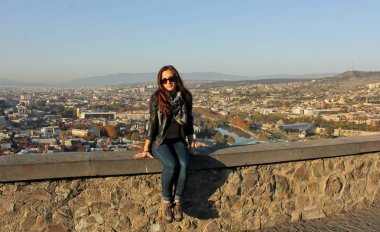 Gezgin kız taş duvarda oturan siyah deri ceket giyiyor. Panoramik, arka plan, eski Tiflis, Gürcistan