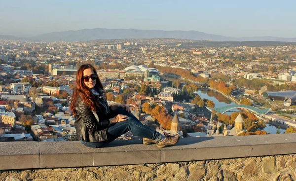 Cestovatel dívka, která nosí černou koženou bundu, sedí na kamenné zdi. Panoramatický pohled ze starých Tbilisi, Gruzie na pozadí — Stock fotografie