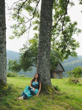 Yaşlı ağacın yanındaki güzel esmer kız. Çimlere basmak. Karpatlar. Dağın yamacında uzun yeşil elbiseli romantik bir kadın..