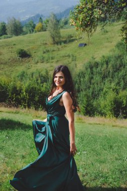 Mutlu, esmer kız çimenlerde dans ediyor. Arka planda Karpatlar var. Dağda uzun, ipek, yeşil elbiseli romantik bir kadın..