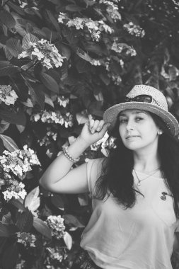 Çalı çiçeklerinin yanında hasır şapkalı güzel bir kız portresi. Bir kadın şapkasını tutar. Genç bir kız çiçek açan vahşi bir çalılığın yanında duruyor. Açık havada siyah beyaz bir kadın portresi.