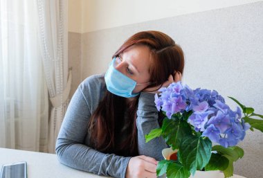 Ev karantinasında tıbbi koruyucu maskeli üzgün bir kadın. Mavi ortanca çiçekli kız. Salgın ruh hali. Koronavirüs salgını sırasında kendini soyutlama. Sağlıklı ve güvenli yaşam konsepti.
