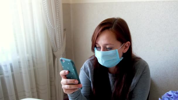悲伤的女人戴着医疗防护面罩在家中隔离 女孩用手机 流行的情绪 在大肠病毒流行期间的自我隔离 女人通过智能手机交流 — 图库视频影像