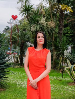 Moda, esmer model, kırmızı takım elbiseli kadın. Seksi, modern tulumlar. Parkta poz veren bir kız, arka planda avuçlar..