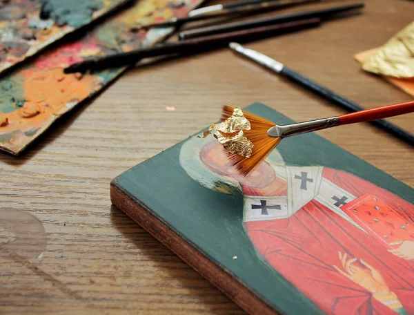Hand with brush of gold leaf. Artist applies gold leaf to the icon. Reproduction of the icon of St. Nicholas. The process of the artist\'s work. Brushes and paints on the background.