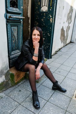 Siyah deri ceketli esmer kadın taş basamakta oturuyor. Rock konsepti. Georgia, Batumi 'deki kız model. Arka plandaki eski yeşil kapı..