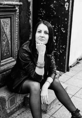 Siyah deri ceketli esmer kadın taş basamakta oturuyor. Rock konsepti. Georgia, Batumi 'deki kız model. Arka plandaki eski kapı. Siyah ve beyaz.