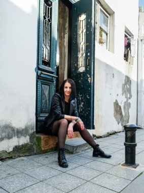 Siyah deri ceketli esmer kadın taş basamakta oturuyor. Rock konsepti. Georgia, Batumi 'deki kız model. Arka plandaki eski yeşil kapı..