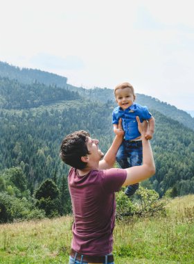 Mutlu genç baba ve küçük oğul dışarıda oynayıp eğleniyor. Arka planda yeşil dağlar var. Yaşam tarzı. Ailenin samimi anları. Babam oğlunu kusuyor. Boş zaman, çocukluk, ebeveynlik ve birlikte hayattan zevk alma kavramı..