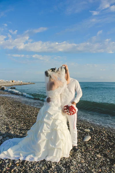 Wedding by the sea. Beautiful couple of newlyweds. Bride with a red bouquet in a wedding dress at the water. Bride and groom kissing on stone beach during sunrise time. Just married in Batumi, Georgia.