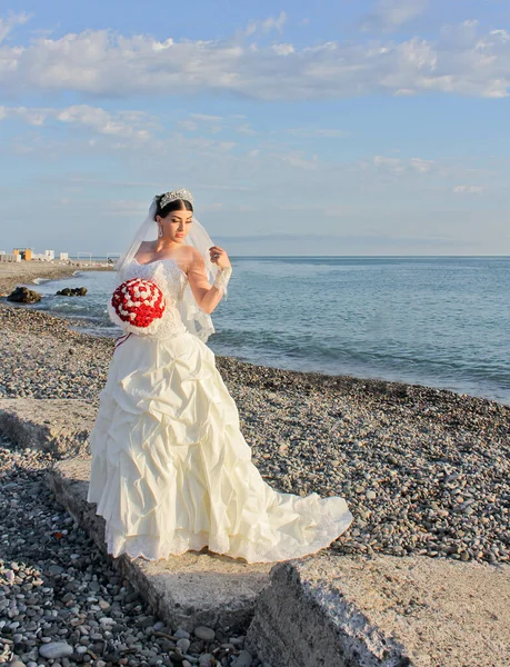 Deniz kenarında düğün. Suda gelinlik giymiş kırmızı buketli güzel bir gelin. Georgia, Batumi 'deki taş plajda Ermeni gelin. Evlilik kavramı, yeni evli.