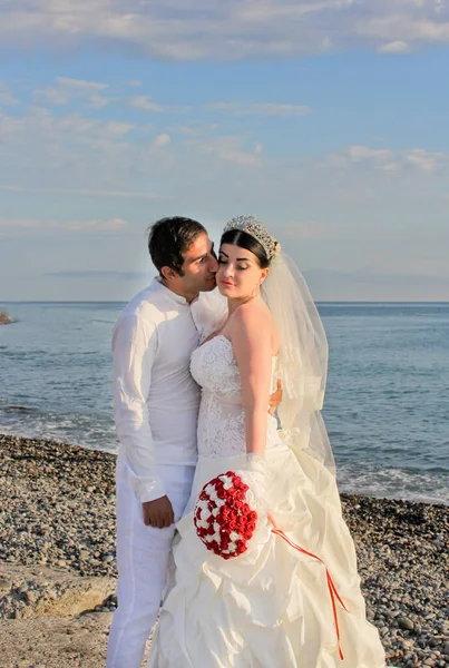 Wedding by the sea. Beautiful couple of newlyweds. Bride with a red bouquet in a wedding dress at the water. The groom kisses the bride on the cheek. Concept marriage, just married.