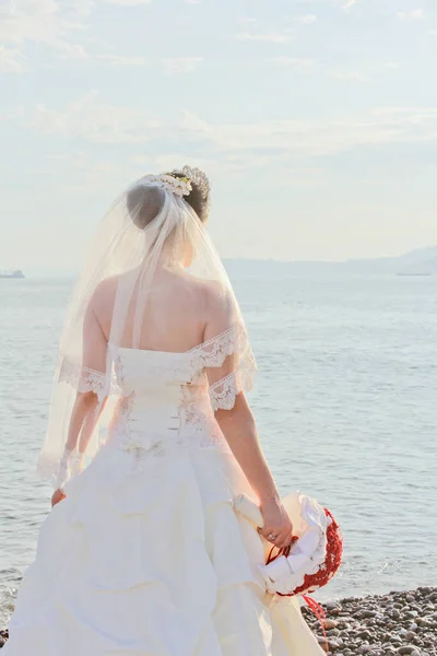 Wedding by the sea. Beautiful bride with a red bouquet. Mountains and sea background. Bride looking at sunrise. Back view. Concept marriage, just married.