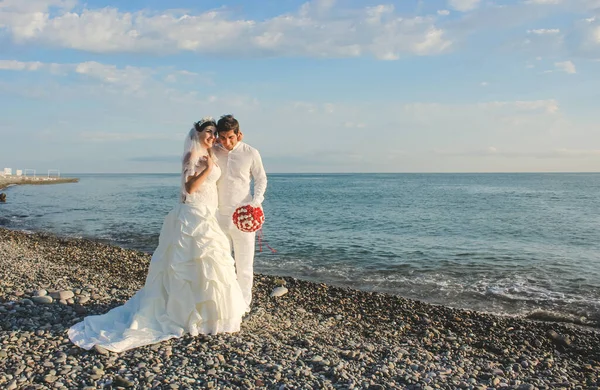 Wedding by the sea. Beautiful couple of newlyweds. Bride with a red bouquet in a wedding dress at the water. Bride and groom smile on stone beach during sunrise time. Just married in Batumi, Georgia.